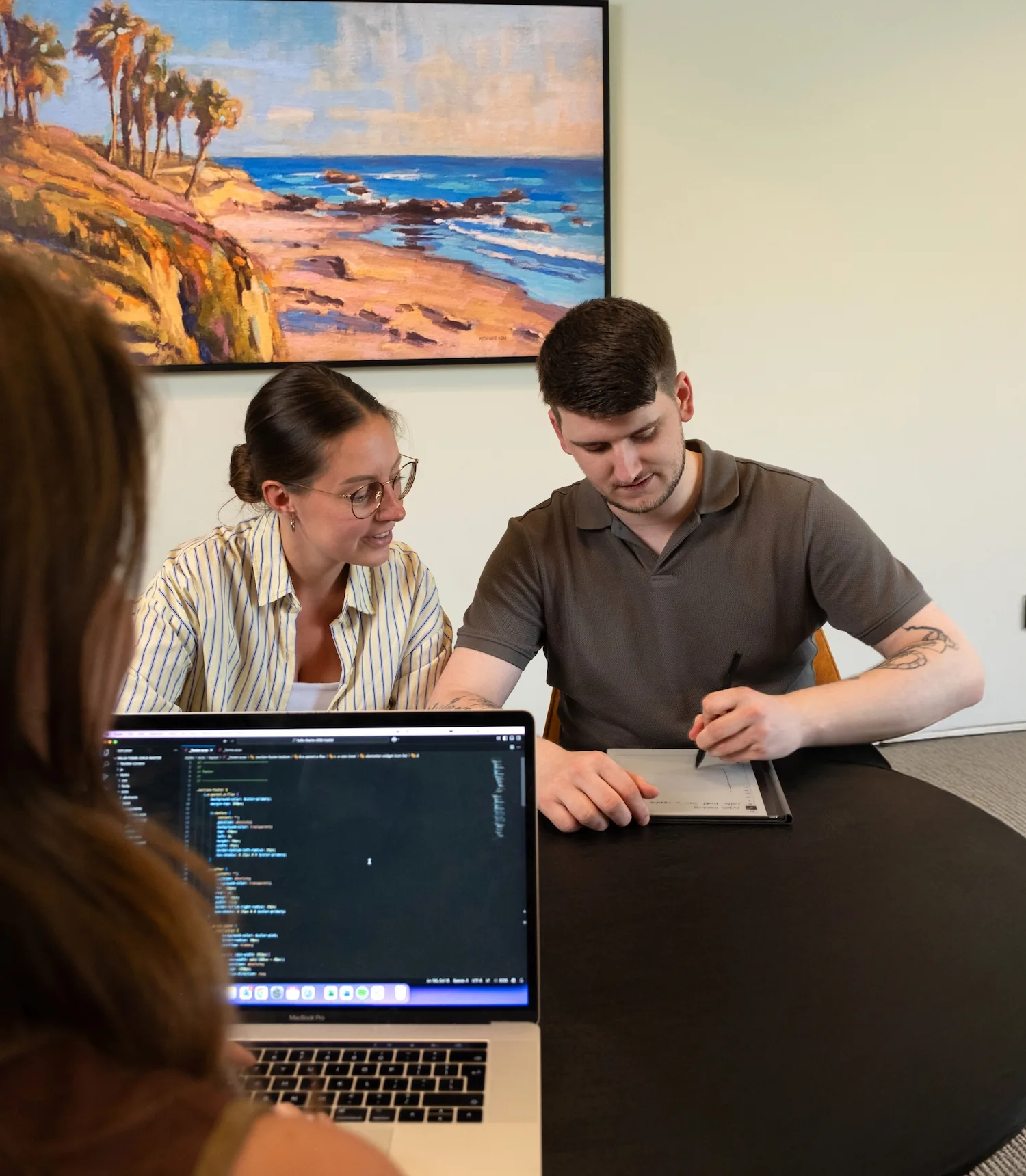 Twee collega’s bespreken een ontwerp op een tekentablet terwijl een derde persoon programmeercode op een laptop bekijkt.