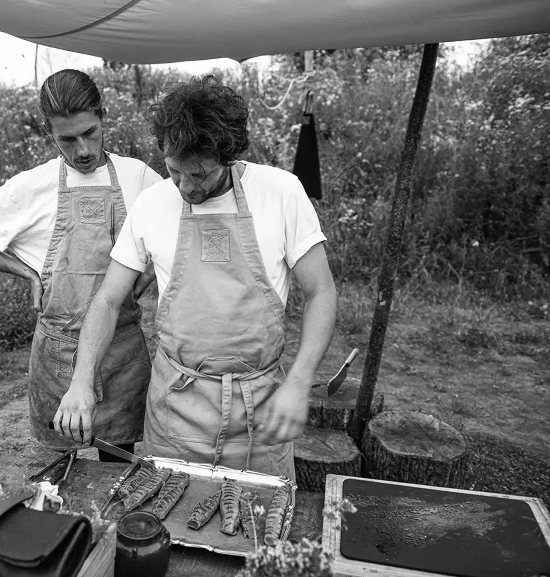 Twee koks in schorten bereiden buiten voedsel op een grillplaat onder een zeil.