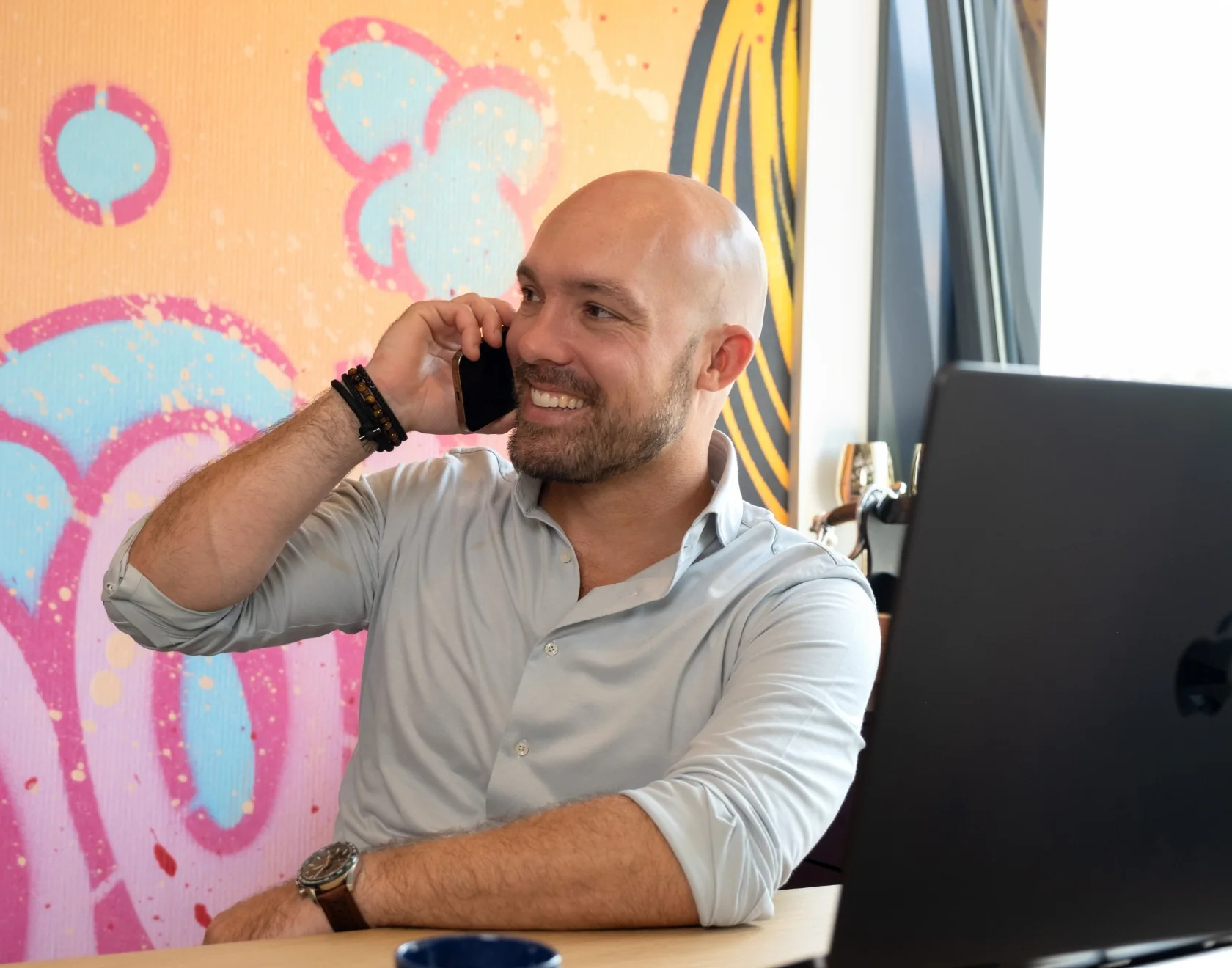 Man aan het telefoneren achter een bureau met laptop en kleurrijke graffiti-achtergrond.