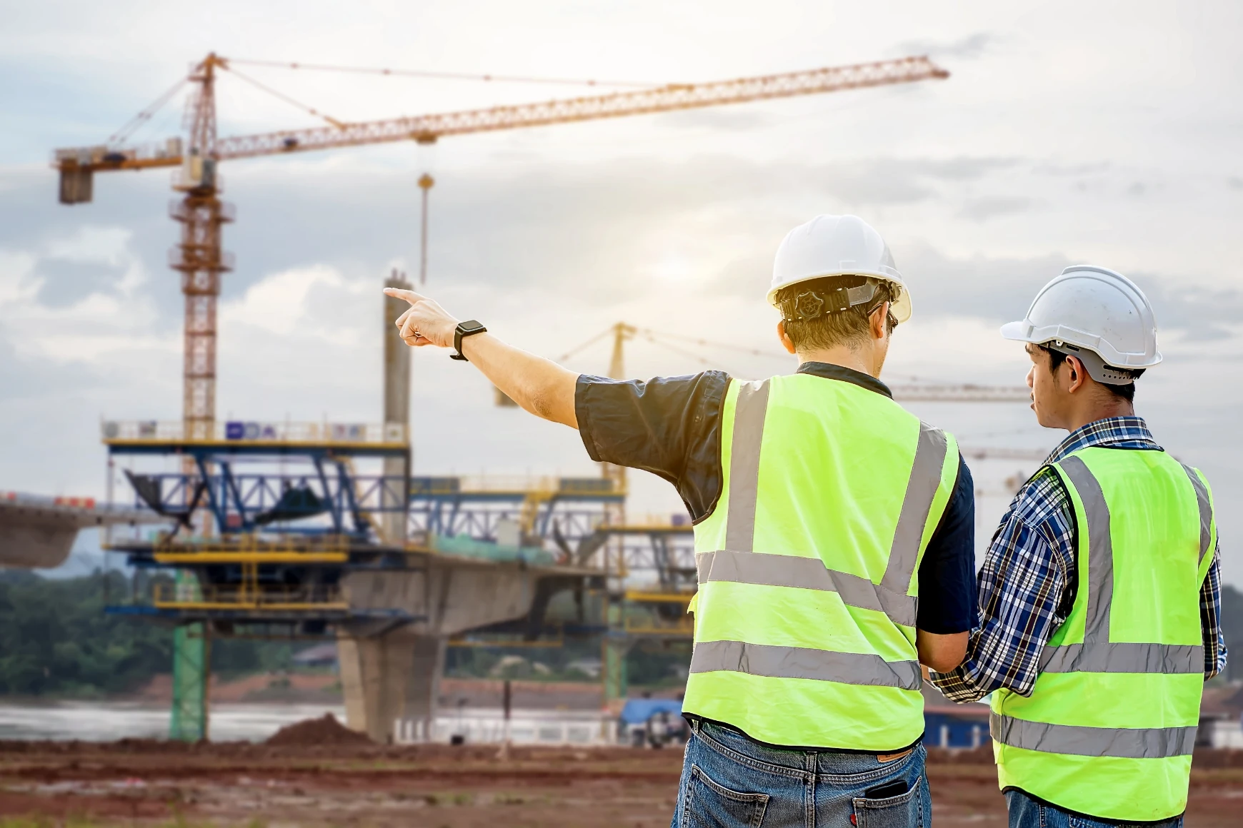 Twee bouwvakkers met veiligheidshelmen en -vesten overleggen bij een grote brugconstructie met hijskranen op de achtergrond.