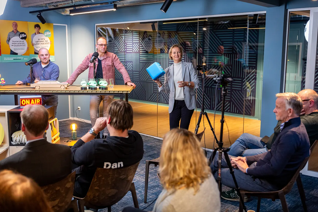 Een live opname van een talkshow met twee presentatoren aan tafel en een vrouw die een blauwe dobbelsteen vasthoudt voor een klein publiek.