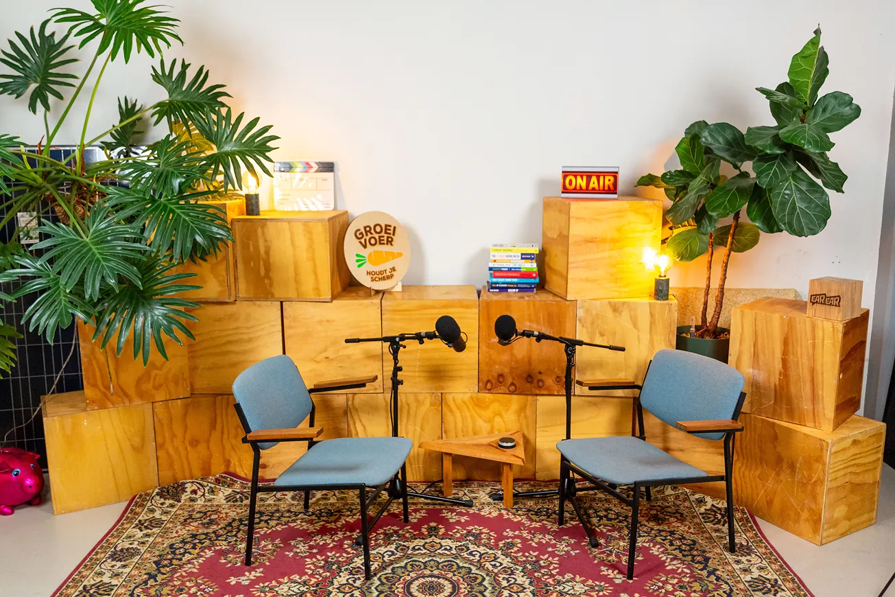 Podcaststudio-opstelling met twee stoelen, microfoons, houten kistdecor, planten en een 