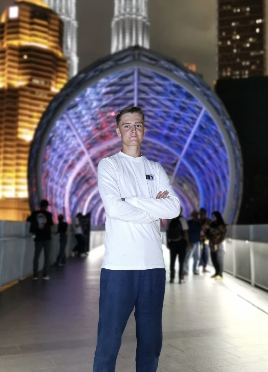 Jonge man poseert met gekruiste armen op een verlicht voetgangersbrug in Kuala Lumpur, met kleurrijk verlichte tunnelstructuur op de achtergrond.